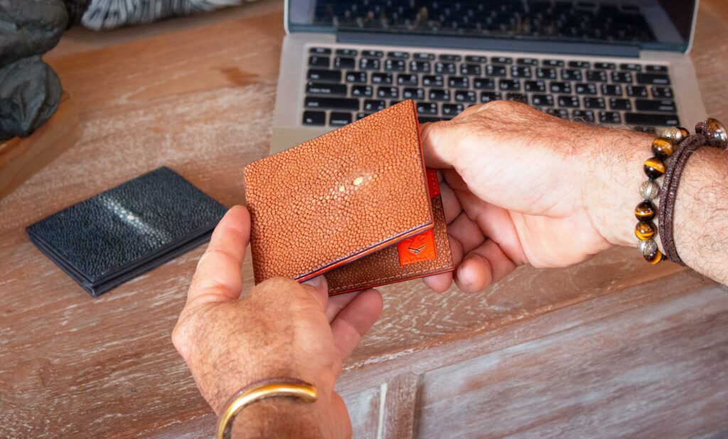 Brown stingray leather bifold wallet by AKNAS with a textured finish, held in hands beside a laptop and black leather wallet.